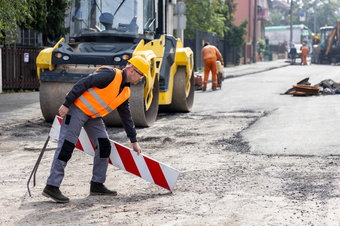 Bauarbeiter stellt Absperrung auf einer Straßenbaustelle auf, während Asphaltarbeiten durchgeführt werden.